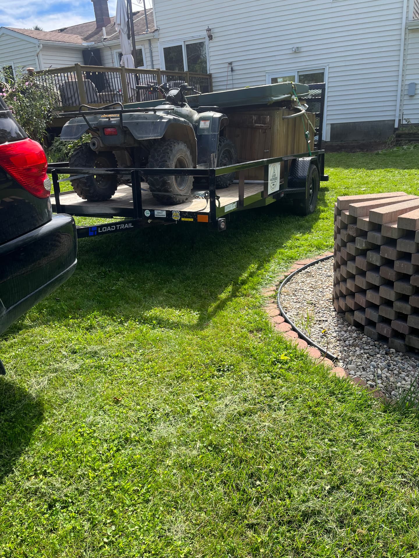 Good Shepherd’s black utility trailer loaded with a four-wheeler and a wooden cabinet, parked on a grassy lawn beside a house